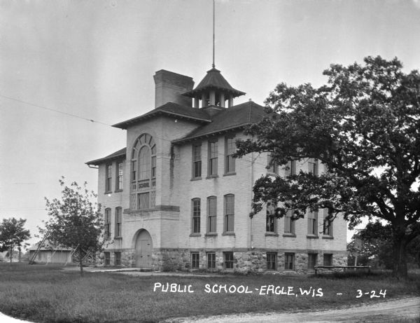 View from the road facing the front and side of the public school built in 1905. A swing set is to the left of the school building. The building has an arched entrance and large arched window above. A bell tower is on the roof.