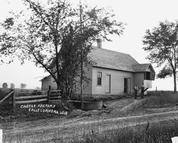 View across dirt road of two men wearing overalls standing outside a rural cheese factory. There is a large, low wooden tub at the driveway entrance, and an overhanging section of the building has an open doorway with a hoist and chains with hooks.