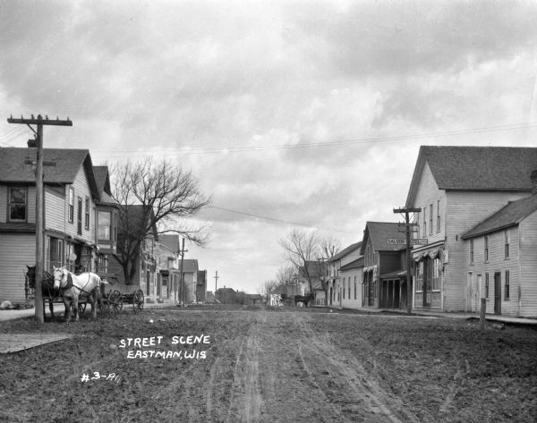 View down an unpaved street. A horse and carriage is tied to a post on the left. A saloon and hotel, and the Park Hotel are located on this street on the right.