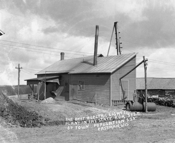 Exterior of the Eastman power plant. Two children pose on a metal cylinder outside the plant. The text on the photograph reads, "The Best electric light plant in the state for size of town - population: 225."