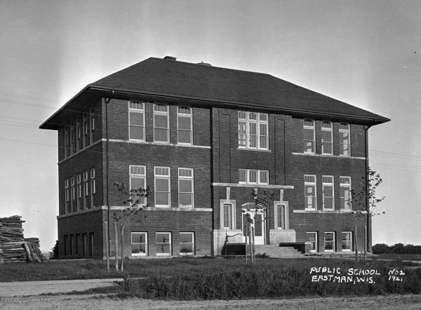 Exterior of the public school in Eastman. On the left is a large stack of logs, and near the entrance to the school is a hand-pump.