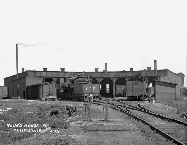 View down side of railroad tracks of two trains entering a Chicago and North Western Railway roundhouse. A man in overalls stands near the tracks.