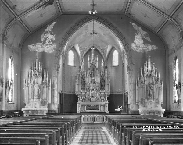 Interior of Saint Patrick's Roman Catholic Church, looking down the aisle of pews towards three altars. The left altar depicts Mary, the right altar depicts Joseph and Jesus. On the wall above flanking the center arch are two murals depicting two angels facing one another.