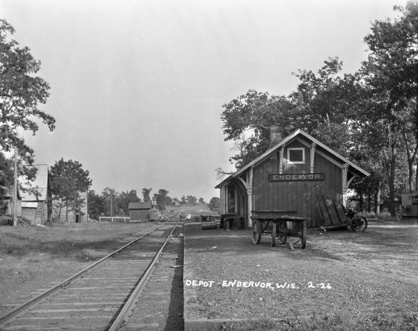 View down railroad tracks of the Wisconsin Central Railway line towards the Endeavor Depot. A man stands near carts, milk cans and barrels near the depot, and barns and other buildings are on the left.
