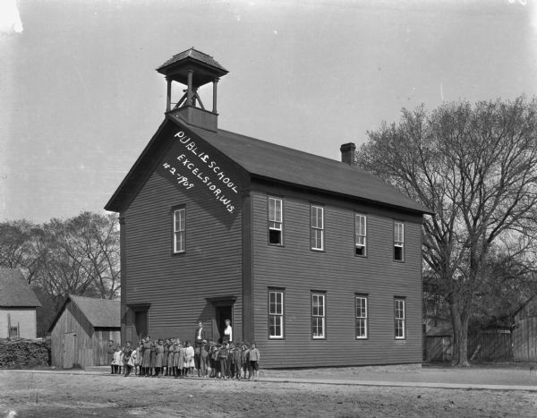View across road of school children and a man and a woman standing in front of the two-story public school. There is a bell tower above the entrance. A number of the children are barefoot.