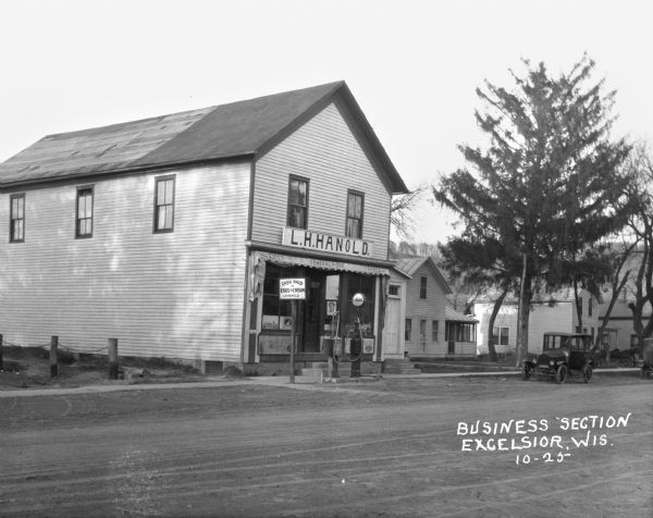 View across road of the exterior of L.H. Hanold, a general merchandise store offering gasoline, tires, tubes, eggs, and cream. In the far background is a tree-lined ridge.