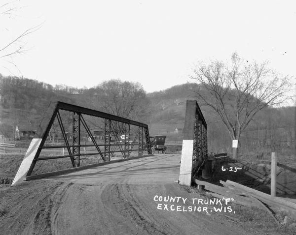 View from road of bridge along County Trunk F. There is an automobile parked on the other side of the bridge. Houses and tree-lined hills are in the background.