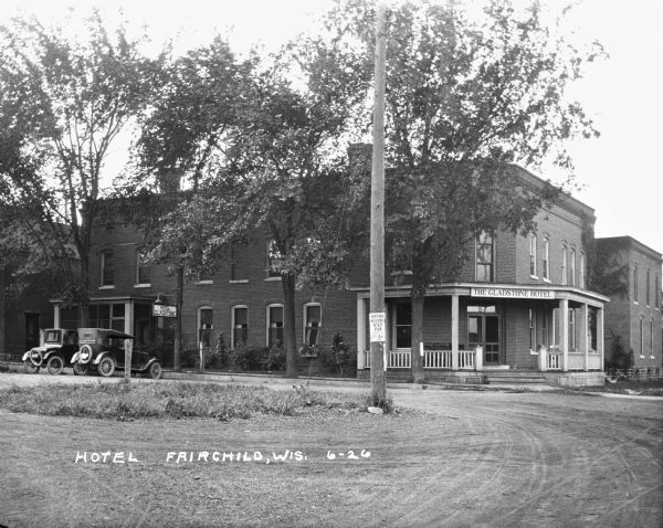 Exterior of the two-story brick Gladstone Hotel. Two cars are parked on the side of the building. An advertisement for the Northern Wisconsin State Fair in Chippawa Falls on September 13 is posted on a utility post in the foreground.