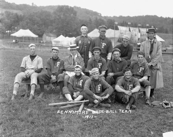 Group portrait of the Fennimore baseball team. In the background there are a number of tents, and large banners and flags fly nearby.
