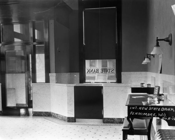 Interior of the newly constructed state bank. Along the wall on the right are writing materials on wall tables, a bench and wall sconces. A window looking out on the street has the words: "State Bank" in reverse. On the left is a glass door leading to a foyer and exterior door.