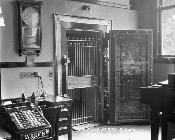Interior of the newly constructed state bank. Featured is a safe, which is open to display the metal gate which has the words: "State Bank of Fennimore" engraved on the center plate. On the left is a Regulator wall clock, and Wales adding machine.