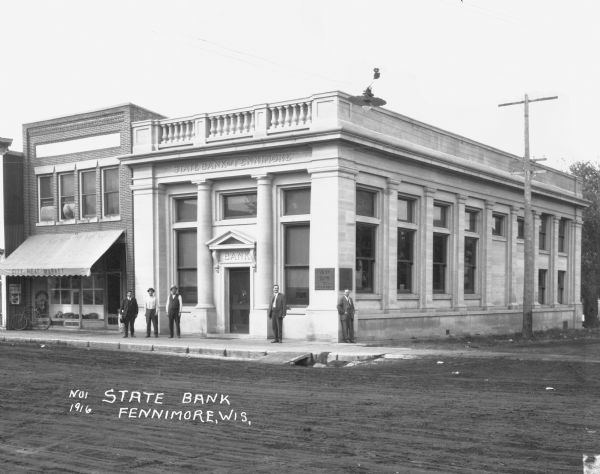 View across street of five men posing on the sidewalk outside the State Bank. Next door to the bank, on the left, is the City Meat Market.