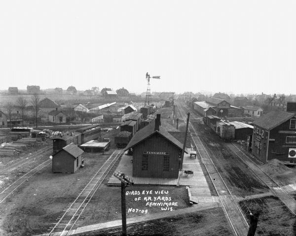 Elevated view of the Fennimore, primarily the train depot. There is a windmill near railroad cars, and dwellings are in the distance.