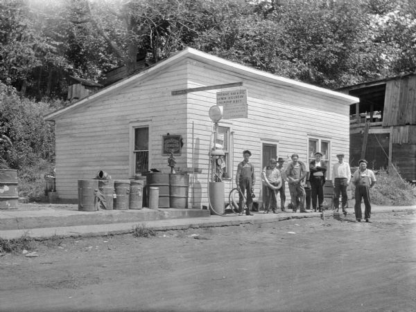 View across road of seven young men posing outside of a variety store. Two men have tires placed over their shoulders. The store's sign advertises: "Tourist, Gas & Oil, Lunch, Ice Cream, Cigars, Pop & Ect." Other buildings are behind the store on a tree and bush-lined hill.