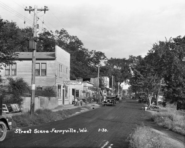 View down street with commercial buildings on the left. There are children on the sidewalk in front of the R.G. Seymour grocery store. Next door is a restaurant, and a few doors down is a sign for "Groceries, Confectionery & [...]its."