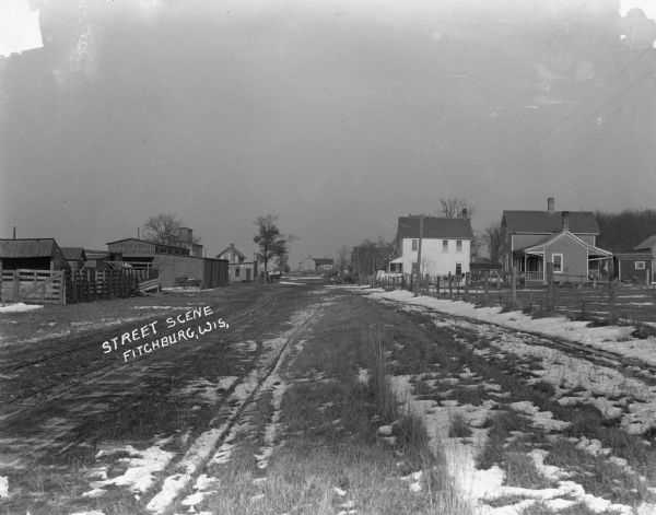 View down dirt road towards the Lappley Bros. Lumber Yard. There are houses on the right and in the far background. There are horses and carriages parked along the edges of the street.