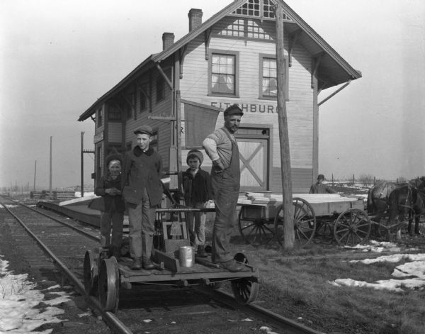 A man and three boys are posing while standing on a handcar at the Fitchburg depot. In the background a man is standing near a horse-drawn wagon.