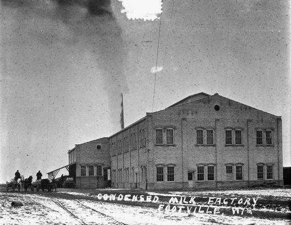 Exterior of a two-story condensed milk factory. Two men are driving horse-drawn wagons on the road on the left. Other men are under a roof covered loading dock in the background with wagons full of milk cans. Heavy smoke is obscuring the large chimney on the factory. Two more people are looking out of an open doorway on the side of the factory.
