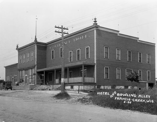 Exterior view from across street of C.J. Hensel hotel and bowling ally. An automobile is parked near the raised platform in front of a storefront with large windows. There is a hand-pump in front of the porch of the hotel.
