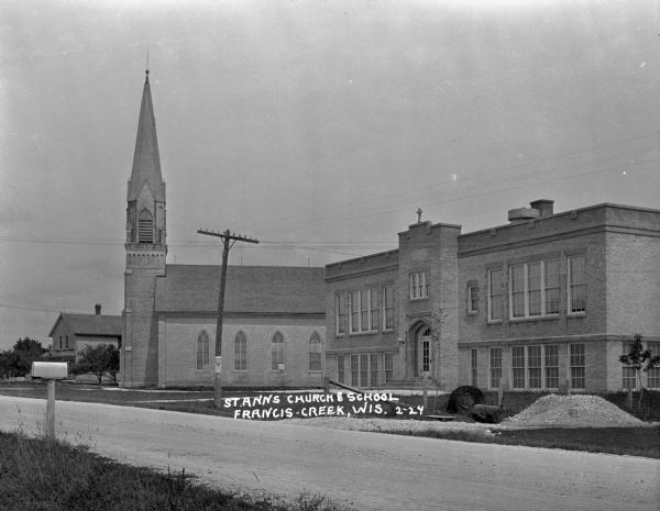 View across Packer Drive of Saint Anne's Catholic Church and Saint Anne Grade School. There is a pile of gravel and other material near a fence in the foreground.