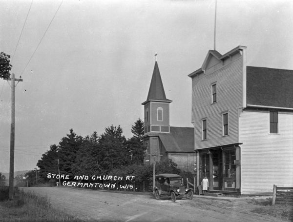 View along an unpaved road. A woman stands at the entrance of a store near a parked car. A church is next door to the store.