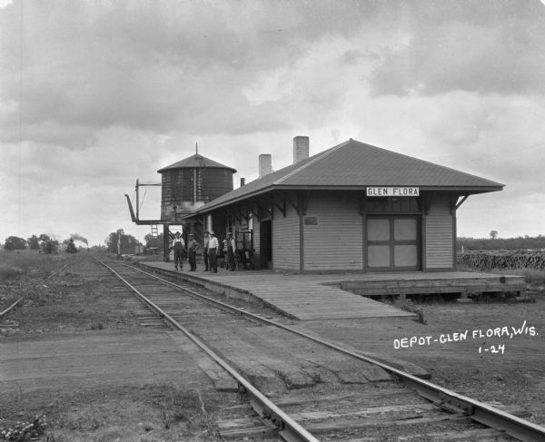 View down railroad tracks of a group of men standing on the platform of the Glen Flora depot waiting for an arriving train which is in the far background.