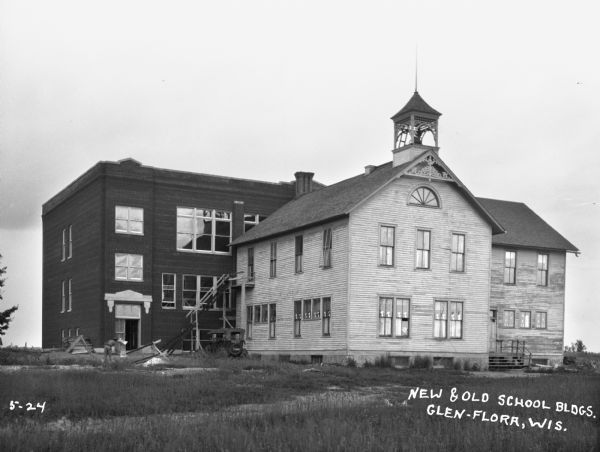 The new school building during construction, next to the old school building, which has a bell tower on the roof. A man works in the yard on the left near a parked automobile.