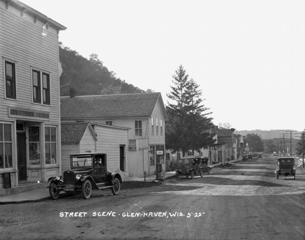 View down a commercial street. A man waits in a car outside H.B. Kness Shop. More automobiles are parked along the curb in the background.