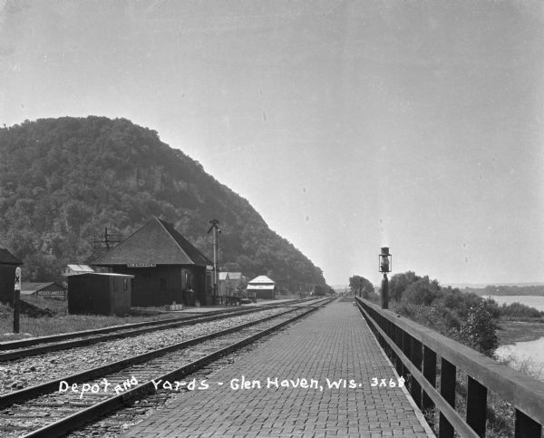 View along a brick platform towards the Glen Haven depot, where two  men stand against the building. Behind the depot is a building with a sign for the "Meuser Lumber Co." On the left is a bluff, on the right is the Mississippi River.