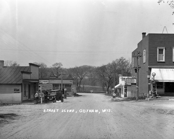 View down middle of road towards two corner shops across from one another. On the left, the store's sign reads, "Hot lunches." Two men pose outside in front of the railroad crossing sign, and behind them is a building that says "Gotham Garage." On the right, the store's sign reads, "Cold meat - cold lunches, cold drinks."