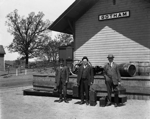 Three men stand, in three-piece suits and hats, in front of the Gotham train depot.