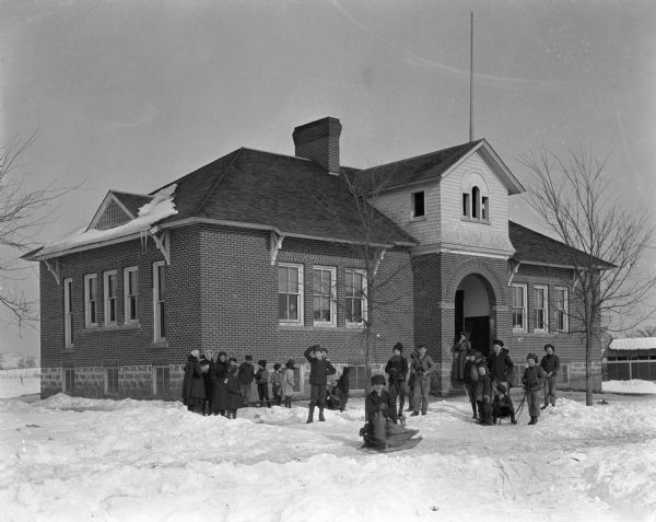 Students in winter attire pose on the snowy lawn of the school. A few of the children have sleds. A woman stands near the arched entrance of the school building.