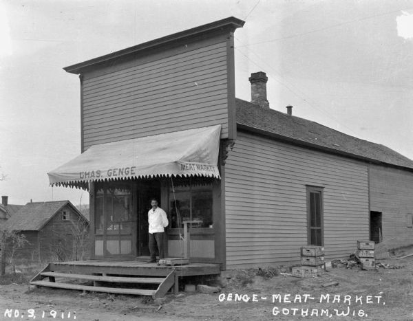 Exterior view of the meat market. A man wearing a butcher's jacket stands on the porch alongside a platform scale.