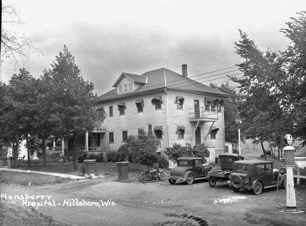 Exterior of the two-story Hansberry Hospital, where three cars and a motorcycle are parked in the adjacent lot.