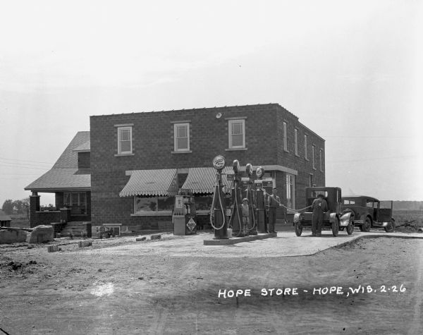 Two men and a child at a filling station and general store in Hope. One man has his back to the camera as he refuels a car.