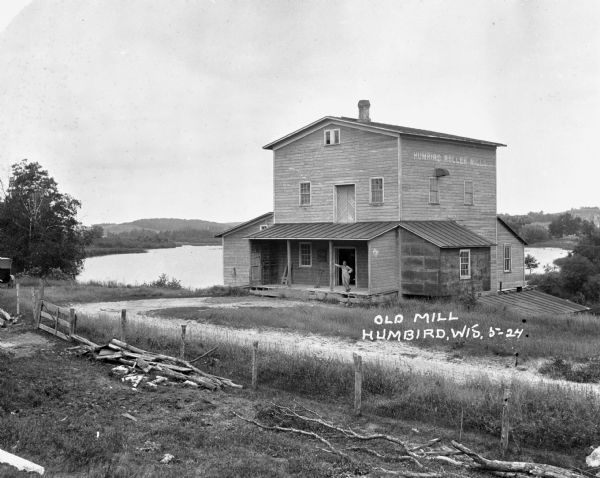 The Humbird Roller Mill surrounded by rural landscape. A man leads on a porch beam as he poses for the picture.