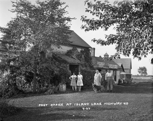 An elderly man, woman, and dog pose near two younger women wearing aprons outside the Island Lake Post Office and Burpee's Resort.