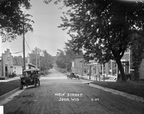 View down road lined with trees and storefronts.  A number of automobiles can be seen parked. The Post Office is on the right.