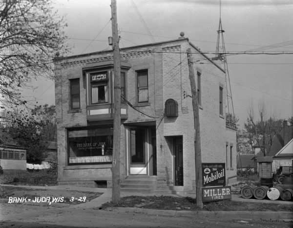 Exterior of the Bank of Juda and an adjacent automotive repair shop. The text painted on the bank window reads, "Safety deposit boxes," "Capital and surplus $25,000.00," and "3% paid on time of deposit." The repair shop has signs advertising Mobiloid and Miller Tires.