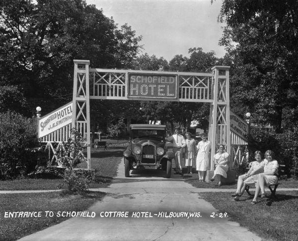A group portrait at the entrance to the Schofield Hotel. A man and five women pose near a parked car at the hotel entrance sign. The sign listed J.A. Smith as the proprietor of the hotel.