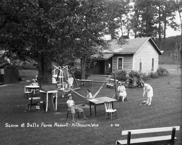 Outdoor scene at the Dells Farm Resort. Five people pose while seated on lawn furniture outside the cabin. Another woman poses as she is about to hit a croquet ball.