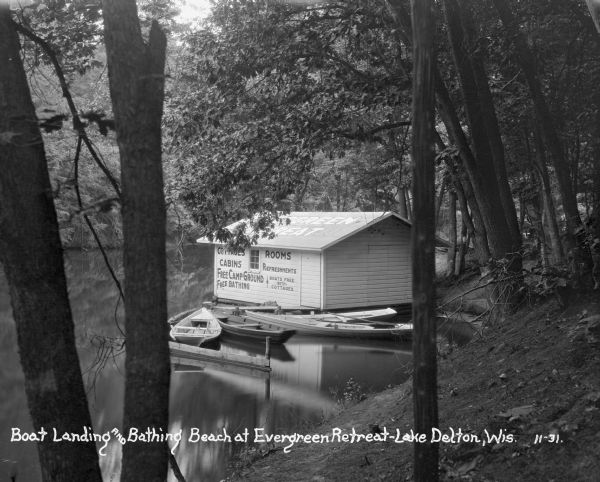 A boathouse and canoe boats along Lake Delton. The boathouse is painted with advertisements that read, "Cottages - Rooms - Cabins - Refreshments - Free Campgrounds - Free Bathing - Boats Free with Cottages."