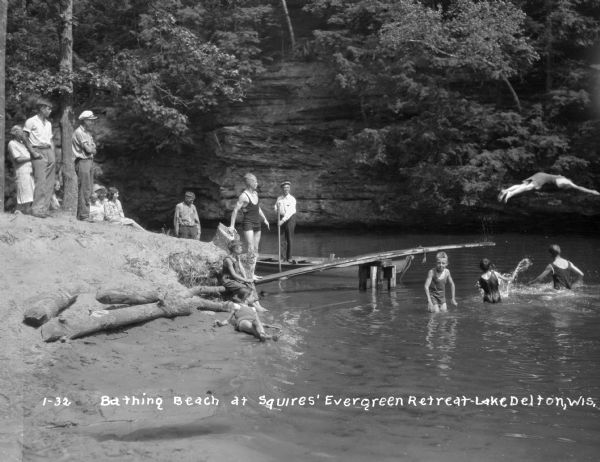 Group of swimmers in bathing suits along the shores of Lake Delton at Squire's Evergreen Retreat. Onlookers observe the activities: children play in the water and a boy is seen diving into the lake.