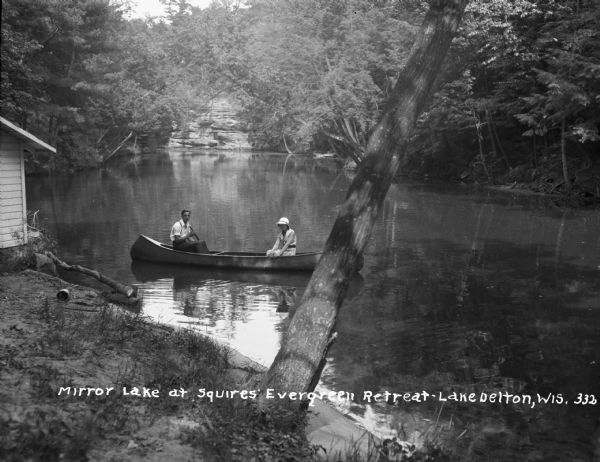 View from shoreline of a man and woman sitting in a canoe on Lake Delton. On the left is part of what appears to be a boathouse.