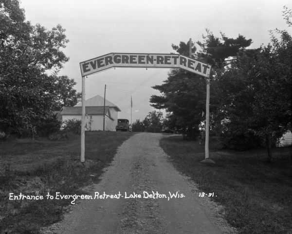 View down driveway of the entrance sign to the Evergreen Retreat. In the background an automobile is parked near buildings and a flag on a flagpole.