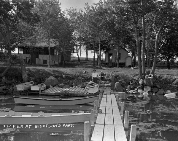 View from on a dock looking at a man and two boys sitting on the rock of the shore. Boats are tied to the dock.