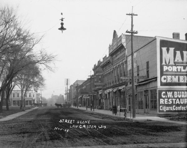 Unpaved main street in Lancaster. The shops along the street include: Smith's restaurant and bakery, Goble and Dyer Furniture, and the Grant County Abstract Office. Two horse and carriages are on the street. A streetlamp hangs over the street from a utility line.