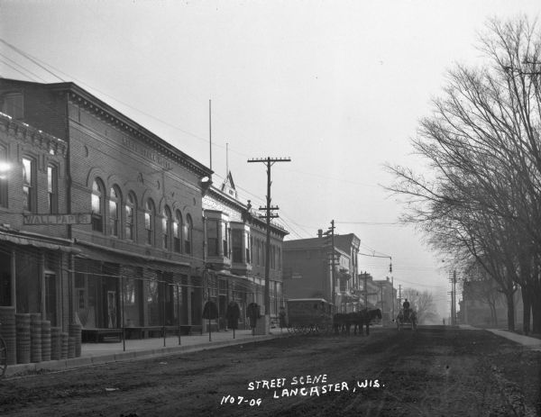 View down commercial street. The shops include: a wallpaper store, Reed Opera House, a clothing store, and a dental office. Two horse-drawn carriages belonging to "Alt and Sons Meats" are parked at the curb.