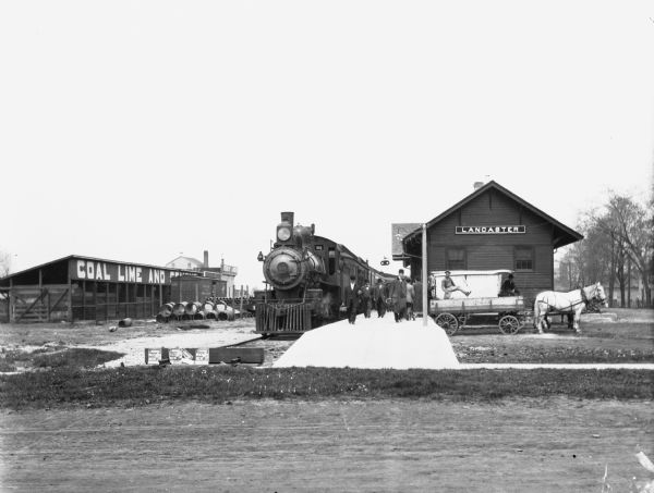View across road of travelers on platform exiting the Lancaster train depot. A man sits on a horse-drawn wagon in front of the depot.