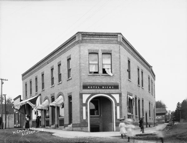 Exterior of the wedge-shaped Hicks Hotel. Lovelace general store is housed on the left side of the building. There are children in the street and on the sidewalk.